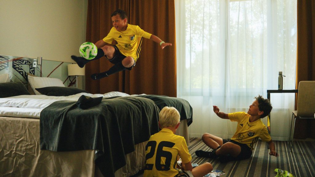 Tre barn i ett hotellrum som leker, de har fotbollskläder och ett barn leker med en fotboll i sängen. De andra spelar kort på golvet.